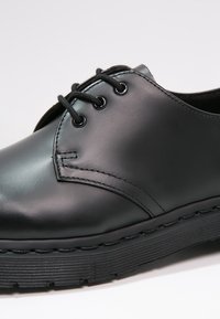 Scarpa in pelle nera con una finitura liscia, punta arrotondata, sei occhielli per i lacci e suola in gomma nera strutturata con scanalature orizzontali.