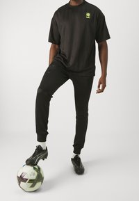 Black athletic set consisting of a short-sleeve shirt with green logo and jogger pants with ribbed cuffs, paired with black soccer cleats on a ball.