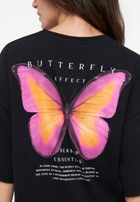 Kobieta nosząca czarną koszulkę z dużą różowo-pomarańczową grafiką motyla oraz napisem "butterfly effect season essentials" na plecach.