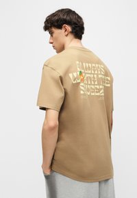 Νεαρός άντρας με κοντά σκούρα μαλλιά, φορεί ένα μπεζ T-shirt με την επιγραφή «Πάντα Αξίζει το Στύψιμο» και ένα μικρό πορτοκαλί σχέδιο στο πίσω μέρος.