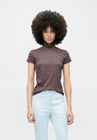 Sieviete ar apjomīgiem cirtainiem matiem, valkājot īs�ās piedurknes topu ar rakstu un gaiši zilas bikses ar augstu vidukli, stāv pret vienkāršu fonu.