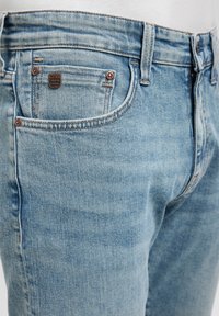 Jeans de mezclilla azul claro con un acabado ligeramente desgastado, cinco bolsillos, acentos de botón de metal y una etiqueta de la marca MAVI en el bolsillo trasero.