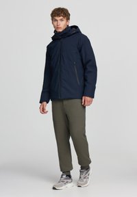 Giubbotto impermeabile blu navy con cappuccio, dotato di tasche laterali con zip, indossato su pantaloni joggers verde oliva e scarpe grigie con suole bianche.