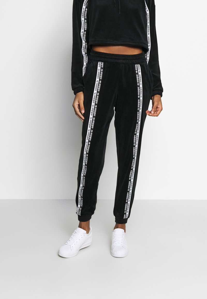 Czarny welurowy dres z białymi paskami po bokach i logo marki, składający się z krótkiej bluzki i dopasowanych spodni ze ściągaczami na nogawkach. Białe sneakersy.