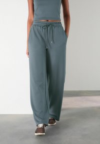 Grijs-groene wijde broek van zacht materiaal, met een elastische tailleband met een trekkoord en zijzakken, gecombineerd met donkere sneakers.