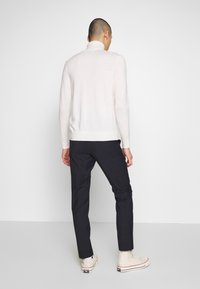 Světle béžový svetr s vysokým límcem, kombinovaný s tmavými kostkovanými kalhotami. Model má na sobě béžové tenisky. Jednoduchý, minimalistický design.