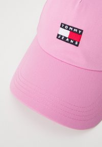 Rozā beisbola cepure, izgatavota no teksturēta auduma, ar taisnstūra Tommy Jeans logotipu, kas satur melnus, baltus un sarkanos elementus.