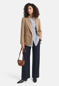 Hellbrauner, doppelt geknöpfter Blazer, weißes und marineblau gestreiftes Hemd, weite marineblaue Hose und eine braune Umhängetasche. Schwarze spitze Schuhe.