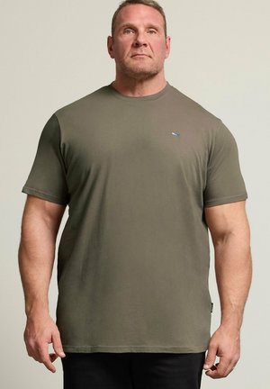 Uomo con capelli corti che indossa una t-shirt verde oliva a maniche corte, in piedi davanti a uno sfondo chiaro e uniforme.
