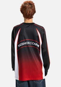 Garās piedurknes melns džersijs ar sarkanu gradientu un baltiem akcentiem. Aizmugurē ir logo "DCSHOECOUSA", izgatavots no gludas, vieglas auduma.
