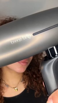 Personne aux cheveux bouclés tenant un pistolet de massage gris étiqueté "3e vitesse", se concentrant sur le panneau latéral et la poignée de l'appareil.