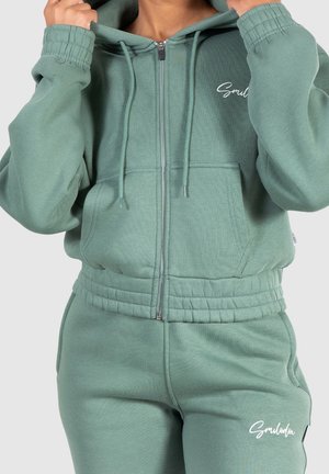 Osoba poprawiająca kaptur, ubrana w zieloną bluzę na zamek i pasujące spodnie dresowe z białymi logo "Smilodox" na piersi i udzie.