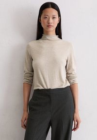 Beżowy sweter z golfem wykonany z miękkiego materiału, z długimi rękawami i luźnym fasonem, w połączeniu z ciemnymi spodniami w prążki.