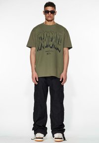 Olivenfarget oversized t-skjorte med fet svart grafisk tekst, parret med svarte cargo-bukser. Modellen har på seg mørke solbriller og joggesko.