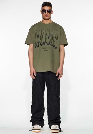 Olivenfarget oversized t-skjorte med fet svart grafisk tekst, parret med svarte cargo-bukser. Modellen har på seg mørke solbriller og joggesko.