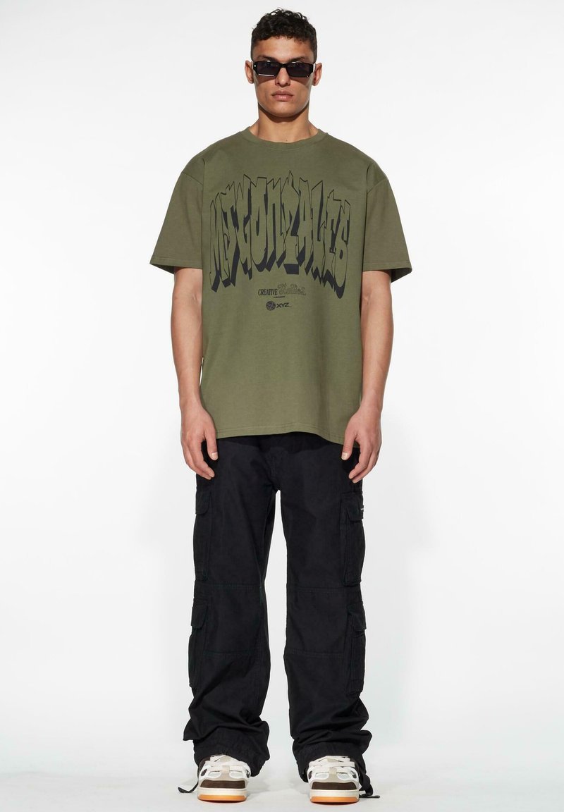 Olivenfarget oversized t-skjorte med fet svart grafisk tekst, parret med svarte cargo-bukser. Modellen har på seg mørke solbriller og joggesko.