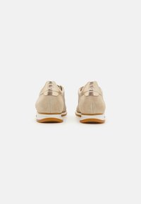 Beige Wildleder-Sportschuhe mit einer strukturierten Oberfläche, kontrastierenden goldenen Akzenten an der Ferse und einer weißen Gummisohle mit einem braunen Boden.