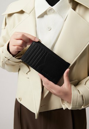 Portafoglio nero dalla texture particolare tenuto in mano, con numerosi scomparti per carte e chiusura con zip, posto su un indumento esterno chiaro e strutturato.
