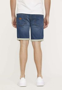 Shorts en denim avec un délavage bleu foncé, ourlet roulé et patch en cuir sur la poche arrière. Portés avec des baskets blanches.