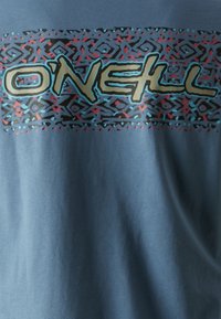 Camiseta de algodón azul con un diseño gráfico que incluye la palabra "O'Neill" en crema, rodeada de patrones geométricos en negro, rojo y azul.