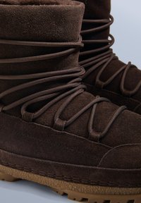 Brązowe zamszowe buty o teksturowanym wykończeniu, z zaokrąglonymi kształtami i wyraźnymi sznurowadłami. Podeszwa gumowa z chropowatym wzorem bieżnika.