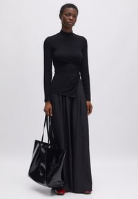 Schwarzes, eng anliegendes Langarmoberteil mit einem Twist-Detail an der Taille, kombiniert mit lockeren, schwarzen, weit geschnittenen Hosen. Glänzende schwarze Tote-Bag und rote High Heels.