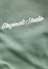 Texte brodé blanc « Originals Studio » sur fond de tissu texturé vert clair.