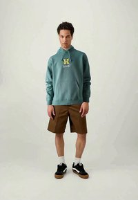 Hooded teal sweatshirt med en framficka och gult HUF-logotyp, ihop med bruna shorts och svarta sneakers, med vita strumpor.