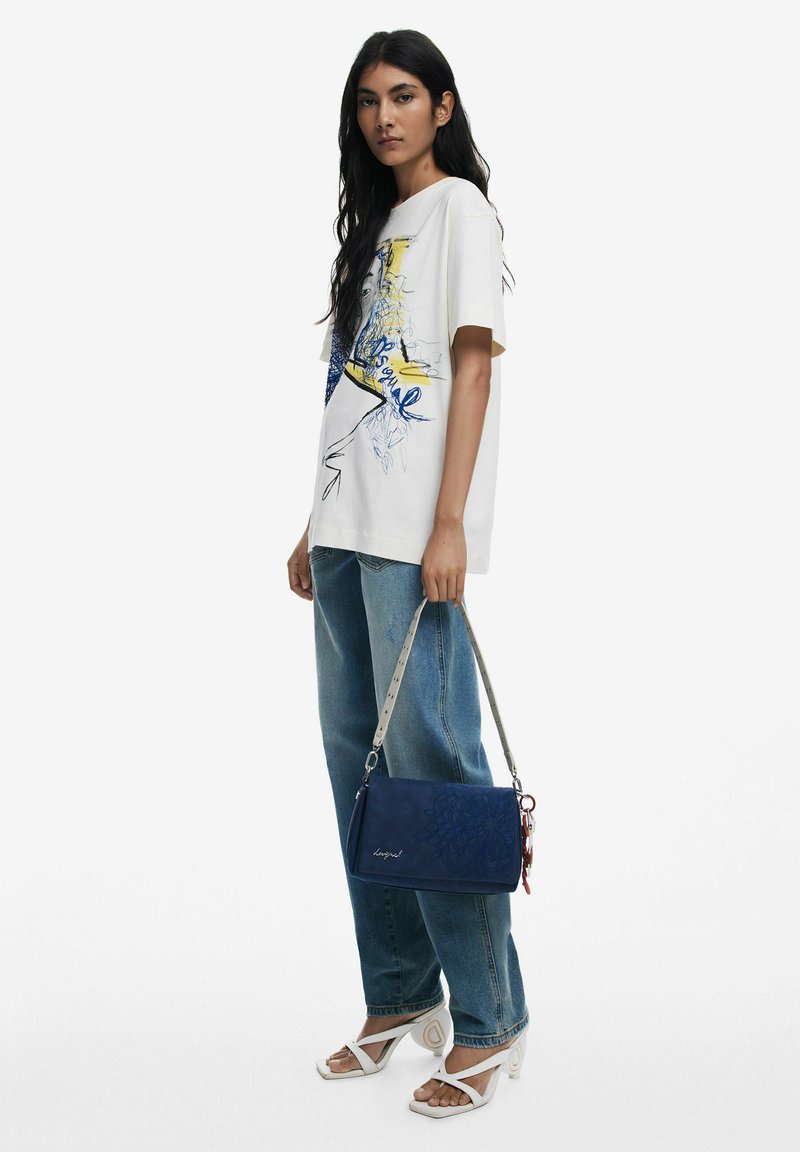 Marineblaue Handtasche mit geprägtem Blumendesign, grauem abnehmbarem Tragegurt und roten Akzenten. Kombiniert mit einem weißen Grafik-T-Shirt und blauen Denim-Jeans.