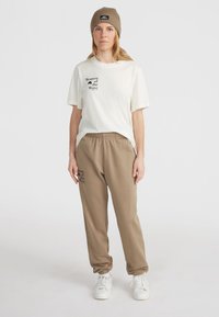 Off-white majica s potiskom "Ženske valov" v črni barvi, v kombinaciji z rjavimi trenirkami, ki imajo prav tako potisk in belimi supergami.