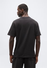 Άν δρας που φοράει απλό μαύρο κοντομάνικο μπλουζάκι και μαύρο παντελόνι, στέκεται με την πλάτη προς το μέρος μας, μπροστά σε ένα απλό φωτεινό φόντο.