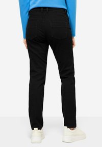 Jeans negros con un diseño ajustado, bolsillos traseros y una cintura de altura media. Llevados con zapatillas blancas sobre un fondo liso.
