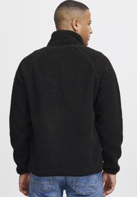 Zwarte fleece pullover met een hoge kraag, gestructureerd oppervlak en raglanmouwen. De stof lijkt zacht en wordt gecombineerd met blauwe jeans.