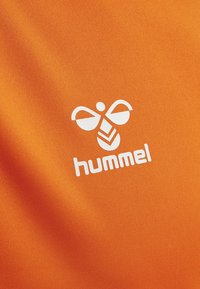 Białe stylizowane logo pszczoły oraz napis „hummel” na jasnopomarańczowym, teksturowanym tle z tkaniny.