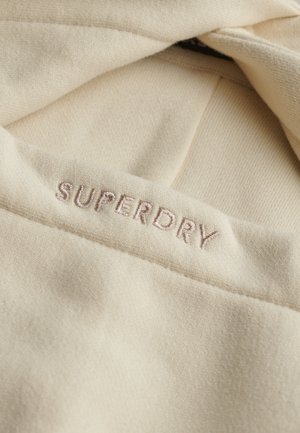 Beige stof met "SUPERDRY" geborduurd in lichtbruine draad op een gevouwen rand, waarbij textuur en stikdetails worden benadrukt.