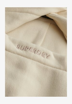 Beige stof med "SUPERDRY" broderet i lys brun tråd på en foldet kant, som fremhæver tekstur og syningsdetaljer.