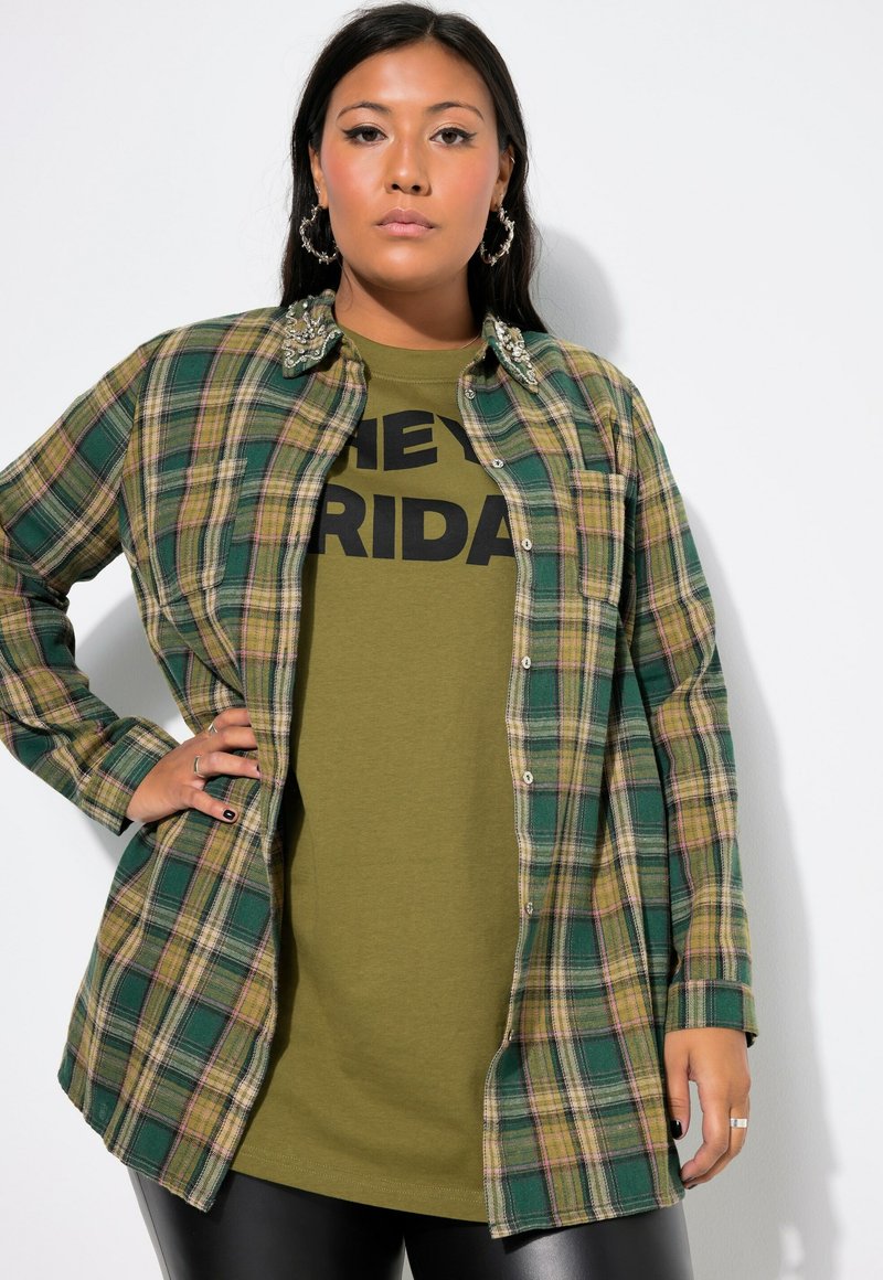 Groene en gele geruite overhemd met knoopsluiting, versierd met kraag, draagt over een olijfgroen t-shirt met vetgedrukte zwarte tekst.