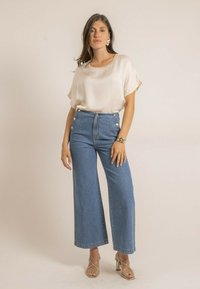 Blouse crème à coupe ample ; jeans bleu à taille haute et jambes larges avec accents de boutons dorés ; sandales à talons dorés avec lanières.
