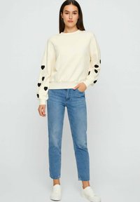 Vrouw in een crèmekleurige trui met zwarte harten op de mouwen, blauwe jeans en witte sneakers, staand tegen een eenvoudige lichte achtergrond.