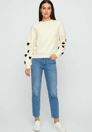 Donna che indossa un maglione crema con cuori neri sulle maniche, jeans blu e sneaker bianche, in piedi contro uno sfondo chiaro e semplice.