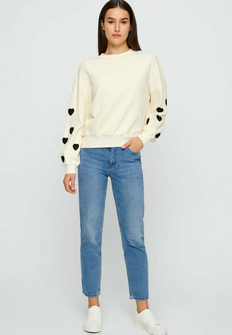 Vrouw in een crèmekleurige trui met zwarte harten op de mouwen, blauwe jeans en witte sneakers, staand tegen een eenvoudige lichte achtergrond.