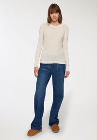 Donna che indossa un maglione crema a maniche lunghe, jeans dritti blu e scarpe slipper marroni, in piedi su uno sfondo bianco semplice.