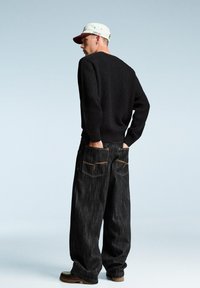 Black knit sweater, loose-fit dark denim jeans with orange stitching and pockets, tan boots, and a light-colored cap with a logo.