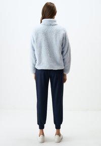 Pullover in pile azzurro chiaro con colletto alto, abbinato a pantaloni jogger blu scuro e sneakers bianche, visto da dietro.