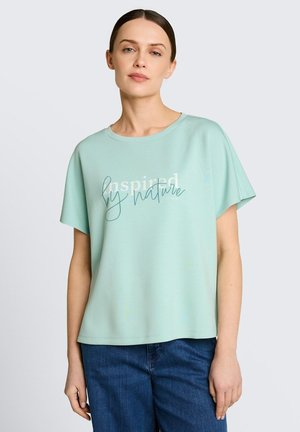 Kobieta o brązowych włosach w miętowo-zielonej koszuli z napisem "Zainspirowana naturą" i niebieskich dżinsach, stojąca na jasnym, jednolitym tle.