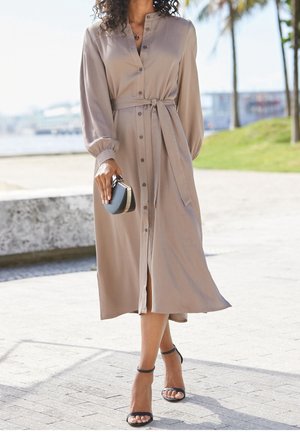 Langes beiges Kleid mit durchgehender Knopfleiste, tailliertem Bindegürtel, ballonförmigen Ärmeln und seitlichem Schlitz, kombiniert mit schwarzen Riemchensandalen und einer kleinen Tasche.