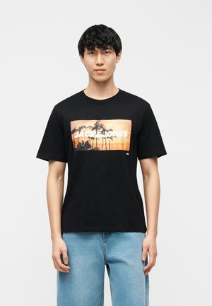 Giovane uomo che indossa una maglietta nera con una grafica di tramonto e palma e jeans azzurri, in piedi davanti a uno sfondo bianco uniforme.