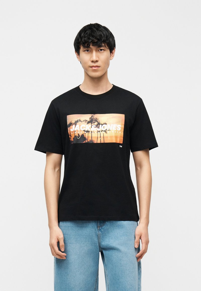 Jeune homme portant un T-shirt noir avec un motif de coucher de soleil et de palmier, et un jean bleu clair, debout devant un fond blanc uni.