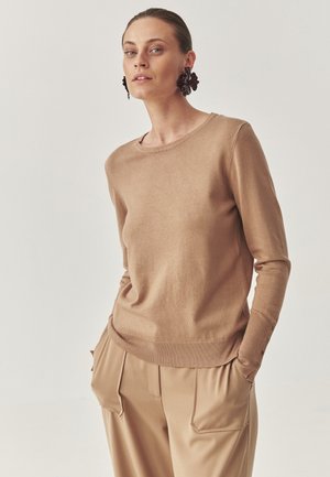Suéter de punto marrón claro con mangas largas, escote redondo, dobladillo acanalado y detalles de botones en los puños, combinado con pantalones beiges.