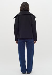 Cappotto di lana nero con ampio colletto e vestibilità rilassata, abbinato a pantaloni di jeans blu a vita alta con tasche laterali, visto da dietro.